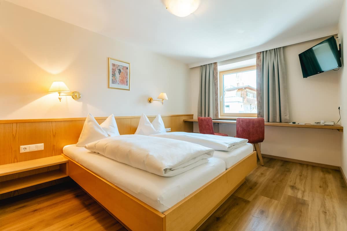 A hotel room with a wooden bed, two bedside tables, a desk with a chair, a flat-screen TV, and a window with curtains.