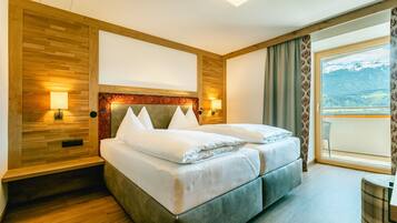 Ein Doppelbett mit Holz-Kopfstück, zwei Nachttischlampen und Blick auf Berge durch ein großes Fenster.