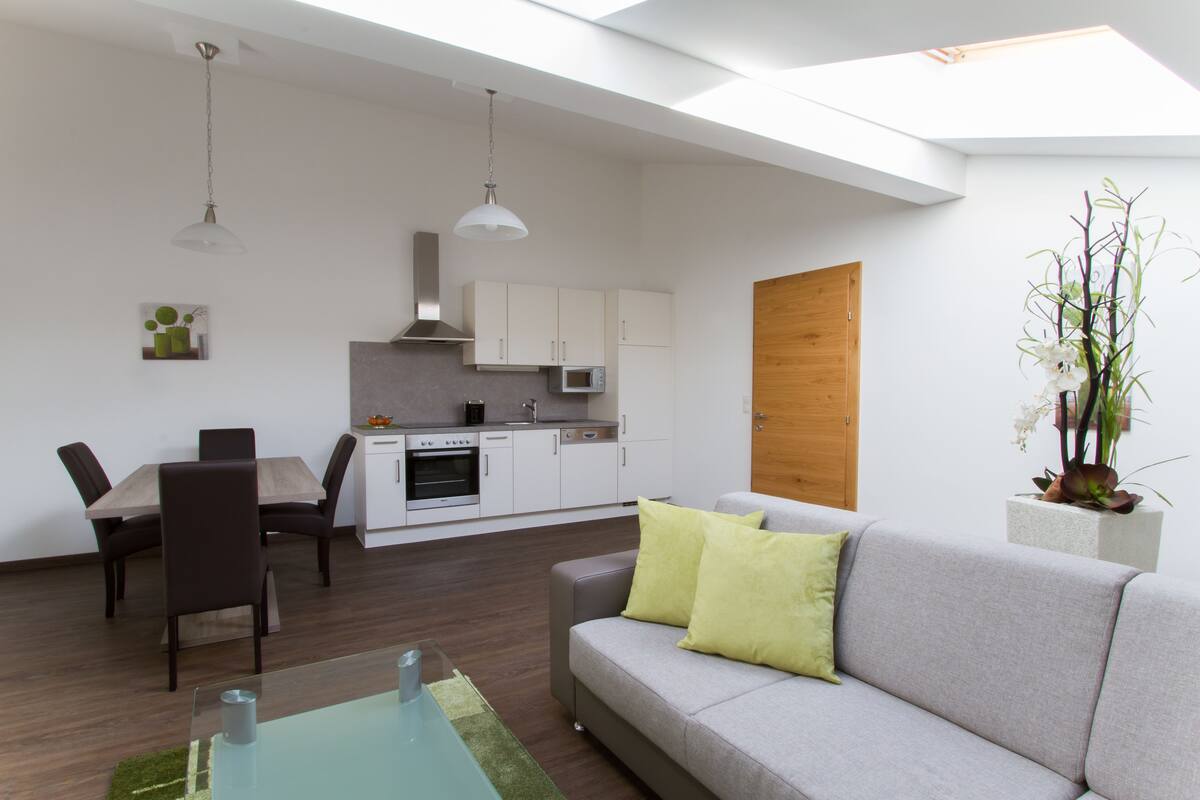 A modern living room with a grey sofa, a dining table with chairs, a kitchen area with white cabinets, and a skylight.