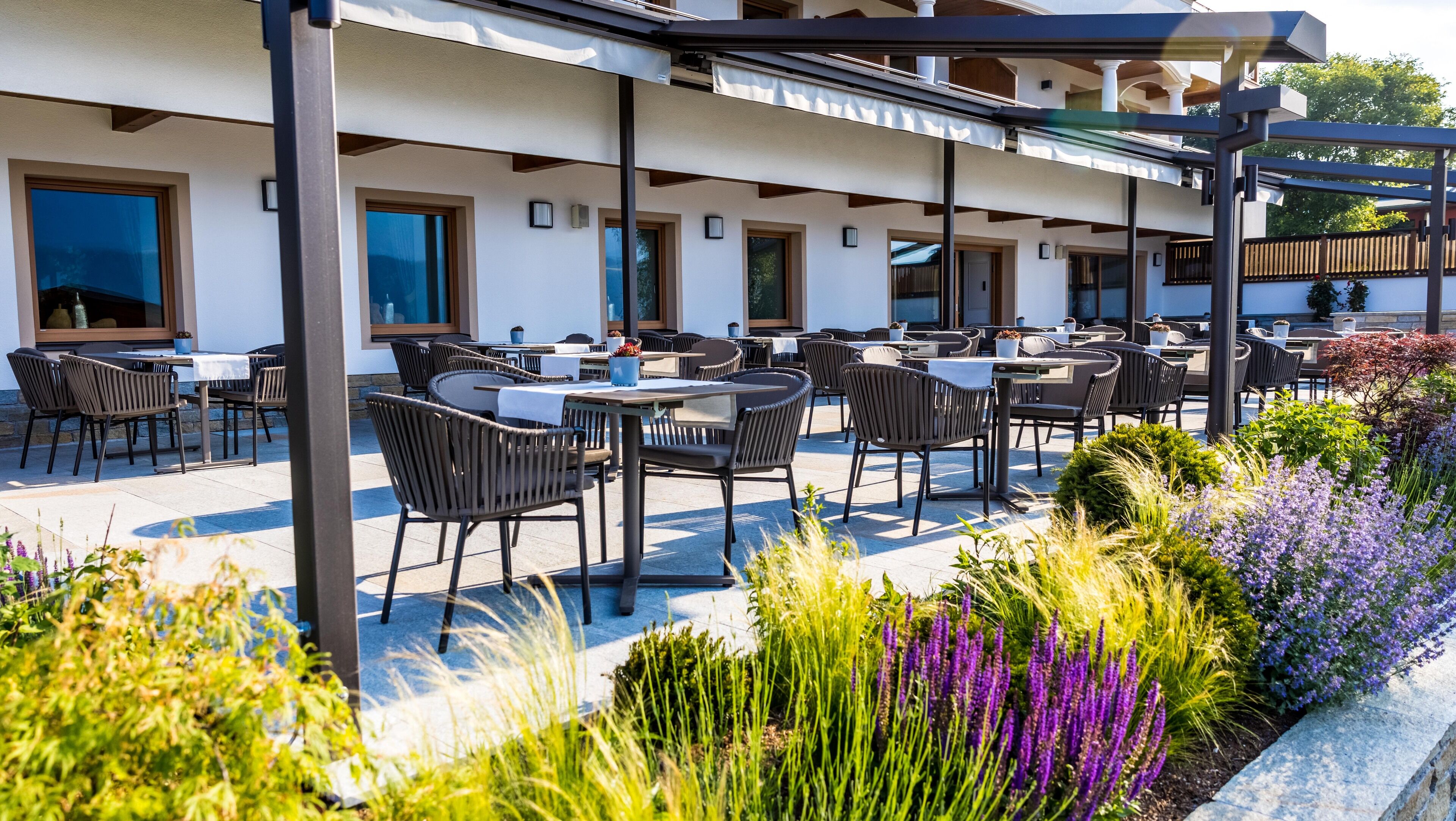 An outdoor patio with multiple tables and chairs, surrounded by potted plants and flowers.
