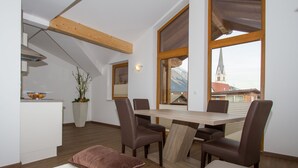 A modern dining area with a wooden table and chairs, a view of a church through large windows, and a kitchen in the background. - Hotel Huberhof (Rum)