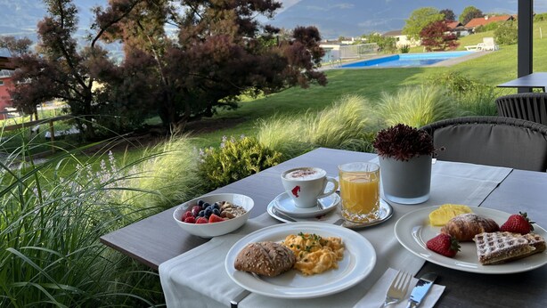 An outdoor dining table with breakfast items, a view of a swimming pool, and mountains in the background. - Hotel Huberhof (Rum)