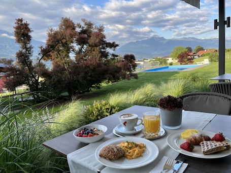 An outdoor dining table with breakfast items, a view of a swimming pool, and mountains in the background.