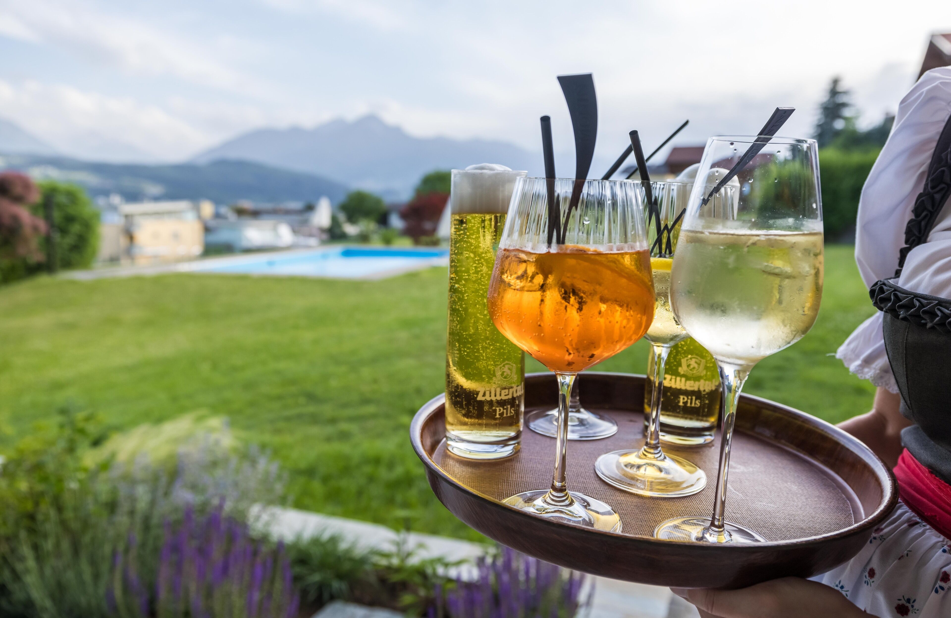 A tray with three glasses of beer and a bottle of beer, set against a backdrop of a grassy area and mountains.