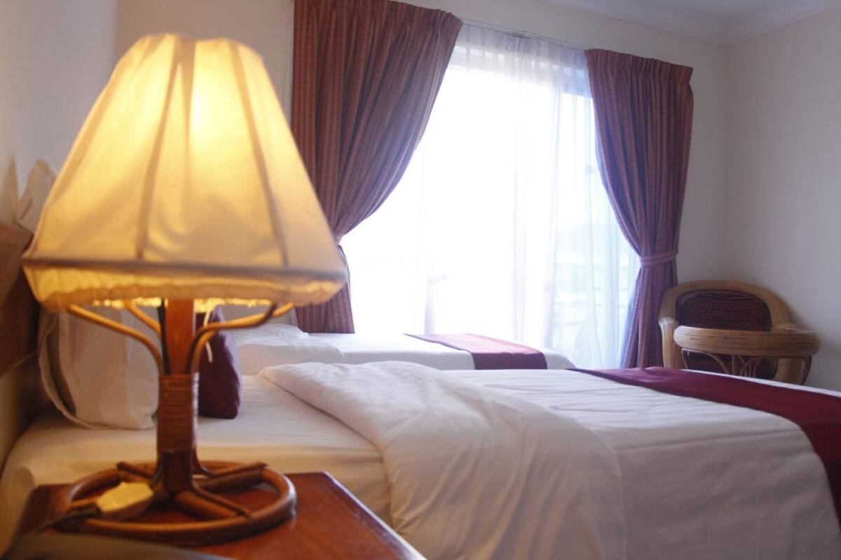 Een hotelkamer met een bed, nachtkastje met lamp, stoel en raam met gordijnen.