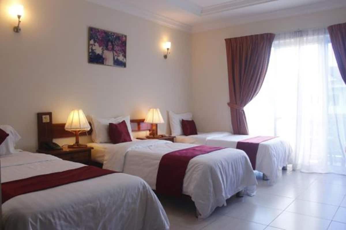 Een hotelkamer met drie enkele bedden, elk voorzien van witte lakens en rode accenten, een nachtkastje met een lamp en een groot raam met gordijnen.