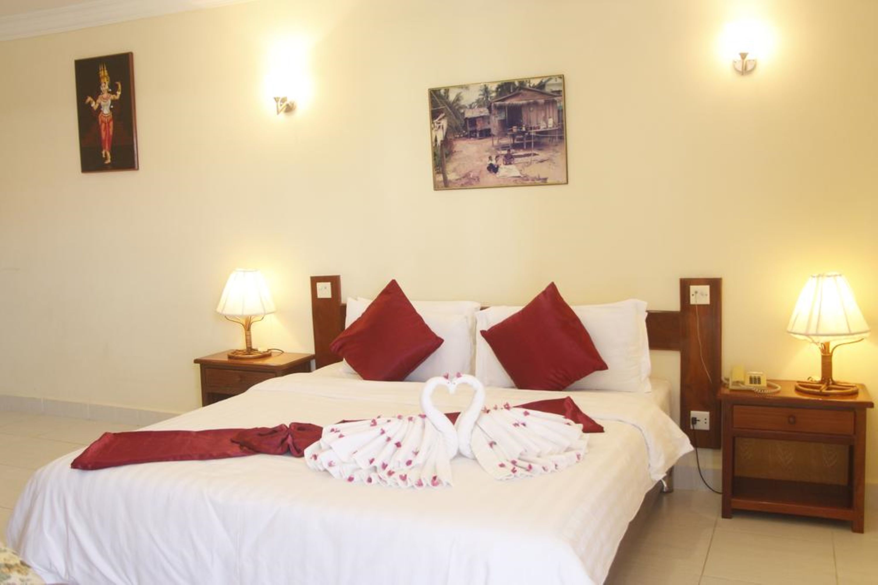 A hotel room with a neatly made bed, two red pillows, a heart-shaped towel arrangement, and a painting on the wall.