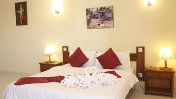 A hotel room with a neatly made bed, two red pillows, a heart-shaped towel arrangement, and a painting on the wall.