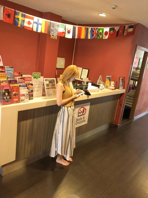 A reception desk with a sign reading "Beds & Dreams" and a variety of informational brochures. Flags are displayed above the desk, and a computer monitor is visible on the counter. - Beds & Dreams Inn @ Clarke Quay (Singapore)