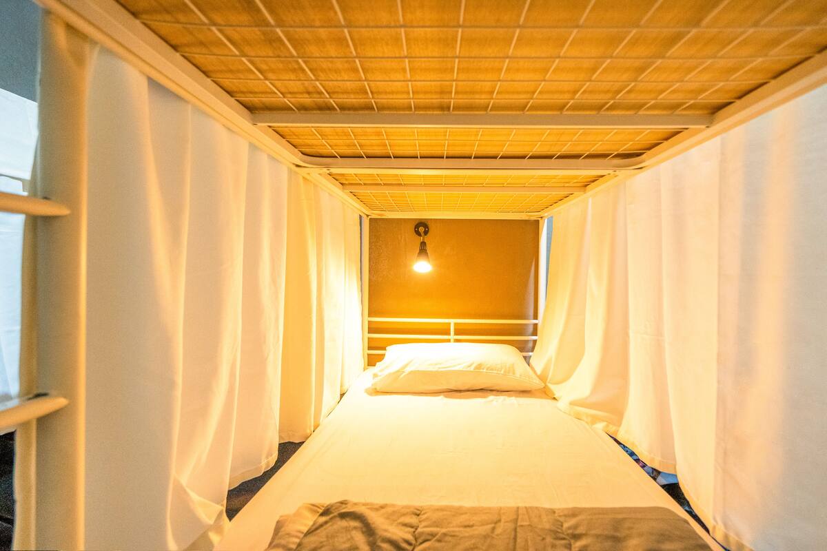 Ein schmaler Raum mit einem Einzelbett, weißen Vorhängen und einer Holzbalkendecke.