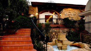 A hotel entrance with a stone facade, a staircase leading up to the entrance, and a small water feature in front. - The Grand Üçel Hotel (Fethiye)