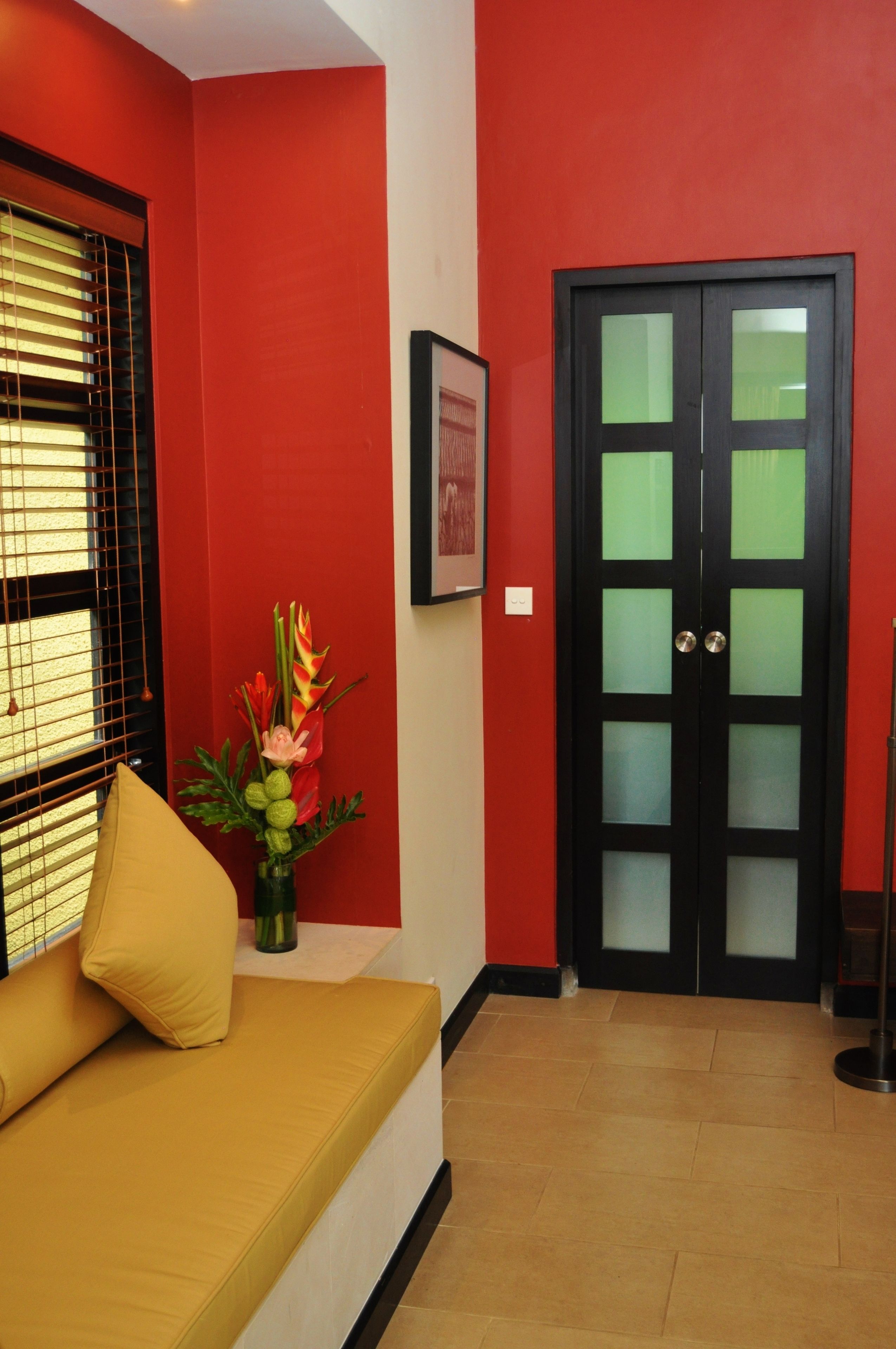 A room with a red wall, a black door with glass panels, a yellow sofa with cushions, and a vase with flowers.