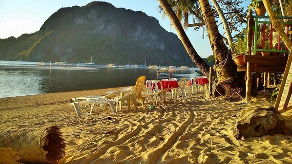 A beach with white plastic chairs and tables, palm trees, and a mountain in the background.