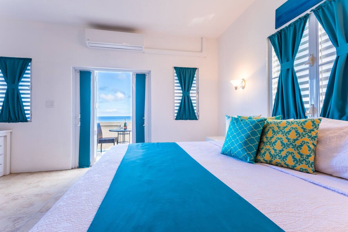 Une chambre avec un grand lit, des rideaux de couleur turquoise et une vue sur la mer, visible par une porte ouverte.
