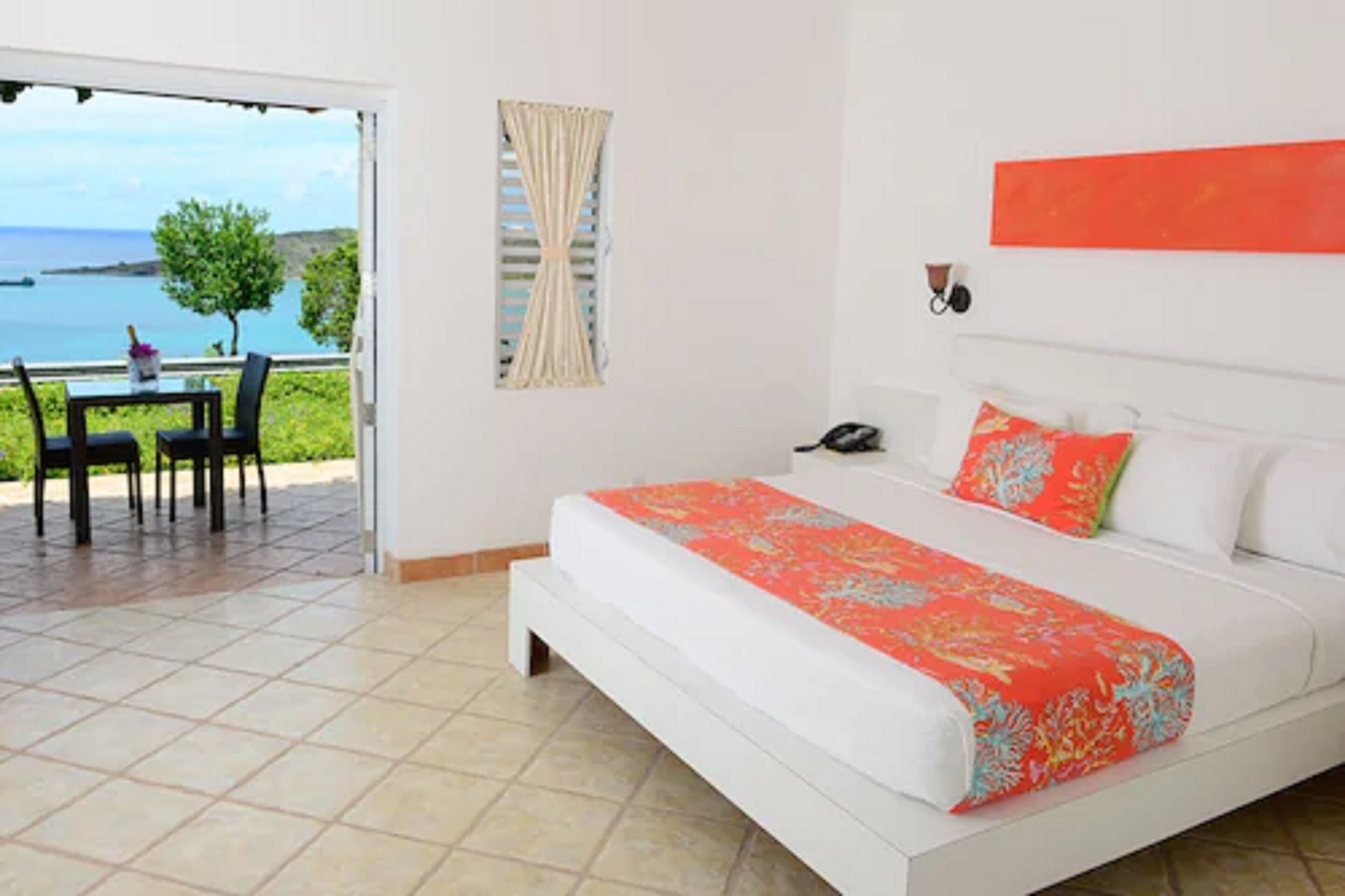 A bedroom with a bed, a nightstand, a telephone, a window with curtains, and a view of the sea.