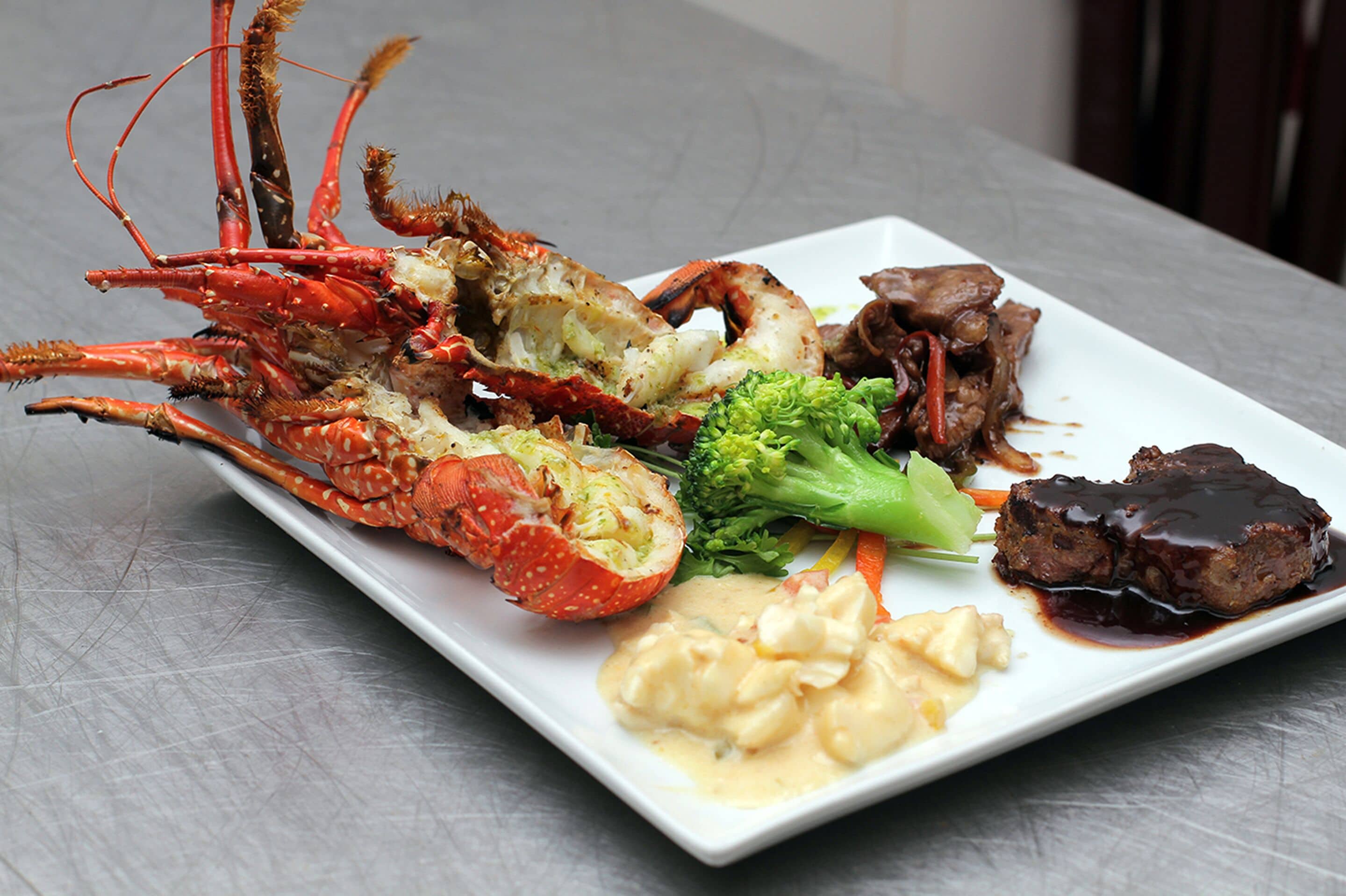 Una bandeja de mariscos con langosta, brócoli, zanahorias y un plato de carne sobre un plato blanco.