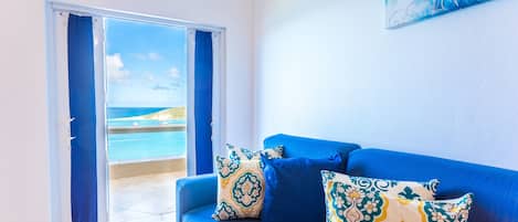 Una habitación con sofá azul, cojines estampados, un cuadro y vista al mar a través de un ventanal.