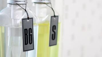Dos botellas de vidrio transparente con dosificador tipo bomba, etiquetadas 'BG' y 'S', sobre una estantería blanca.