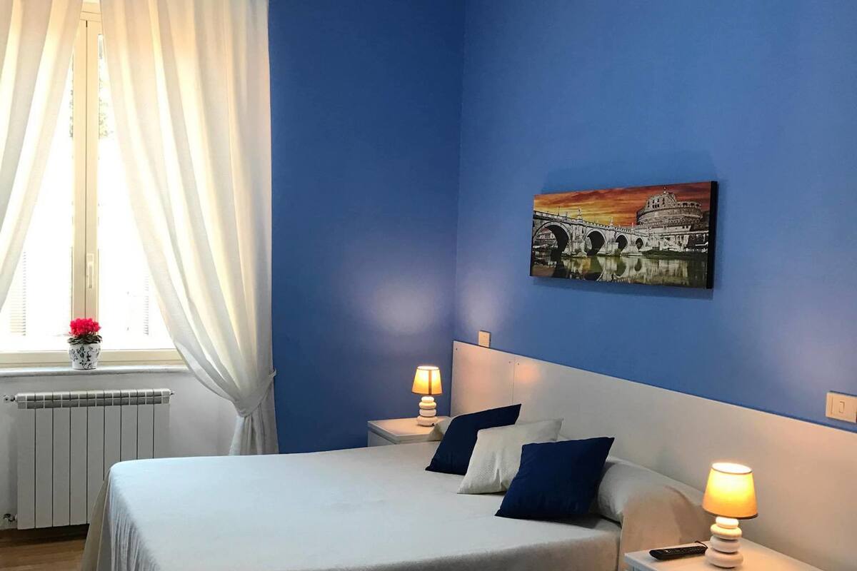 A bedroom with a large bed, two bedside tables with lamps, a window with white curtains, a radiator, and a painting on the wall.