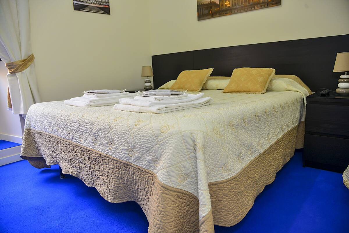 A neatly made bed with a quilted bedspread, two pillows, and folded towels on a blue carpeted floor.