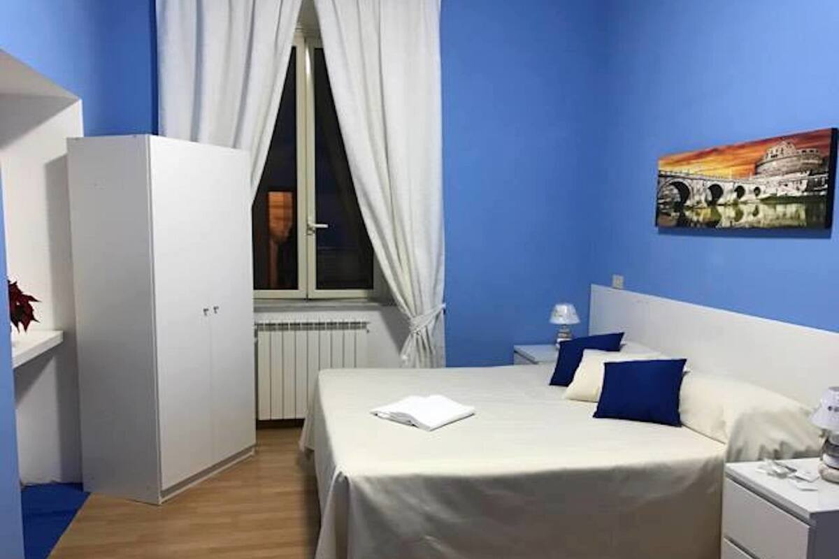 A hotel room with a bed, bedside tables, a nightstand, a window with curtains, and a wall painting.