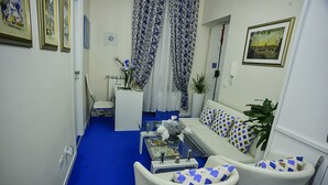 Una sala de estar moderna con alfombra azul, muebles blancos y elementos decorativos como cortinas y cuadros enmarcados.
