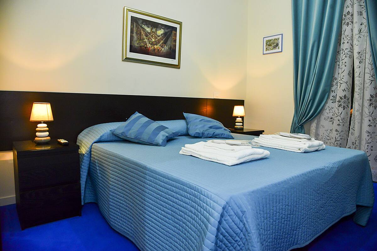 A neatly made bed with blue bedding and pillows, a nightstand with a lamp, and a framed picture on the wall.