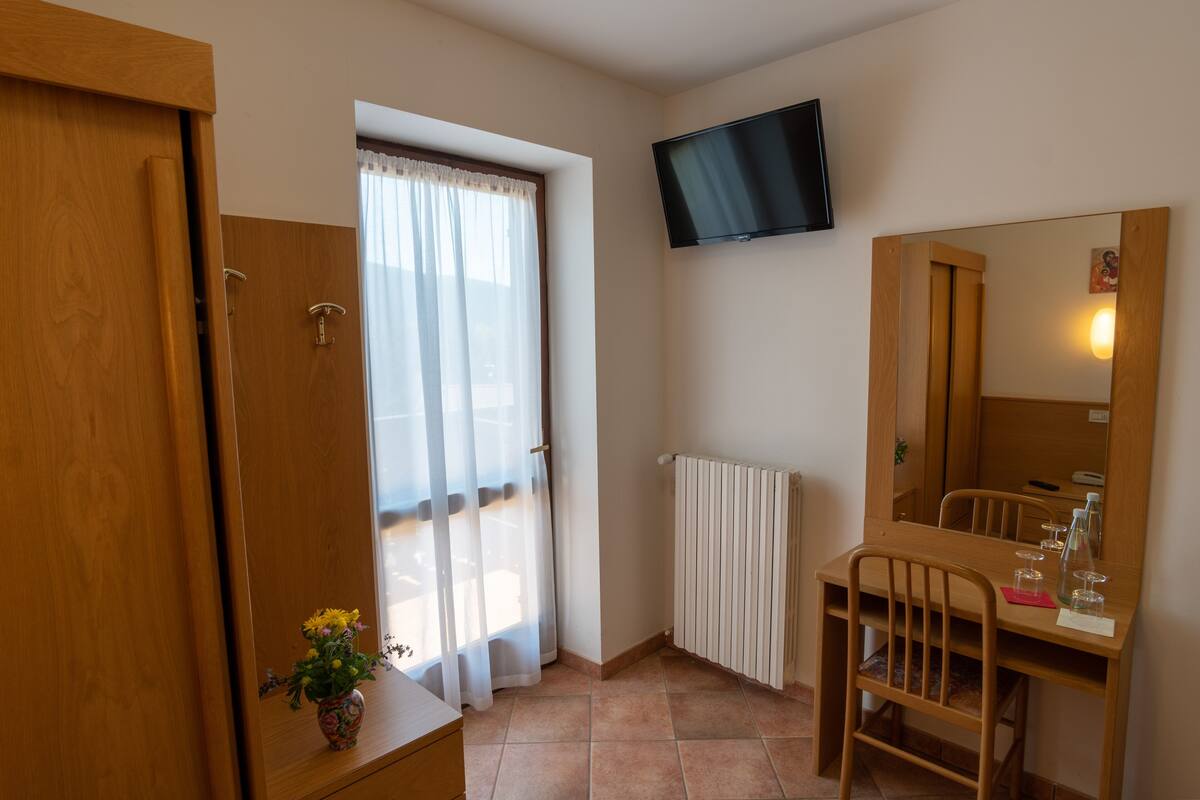 Ein Zimmer mit Holzschrank, Schreibtisch mit Stuhl, Spiegel, Wand-Fernseher und Fenster mit Vorhängen.