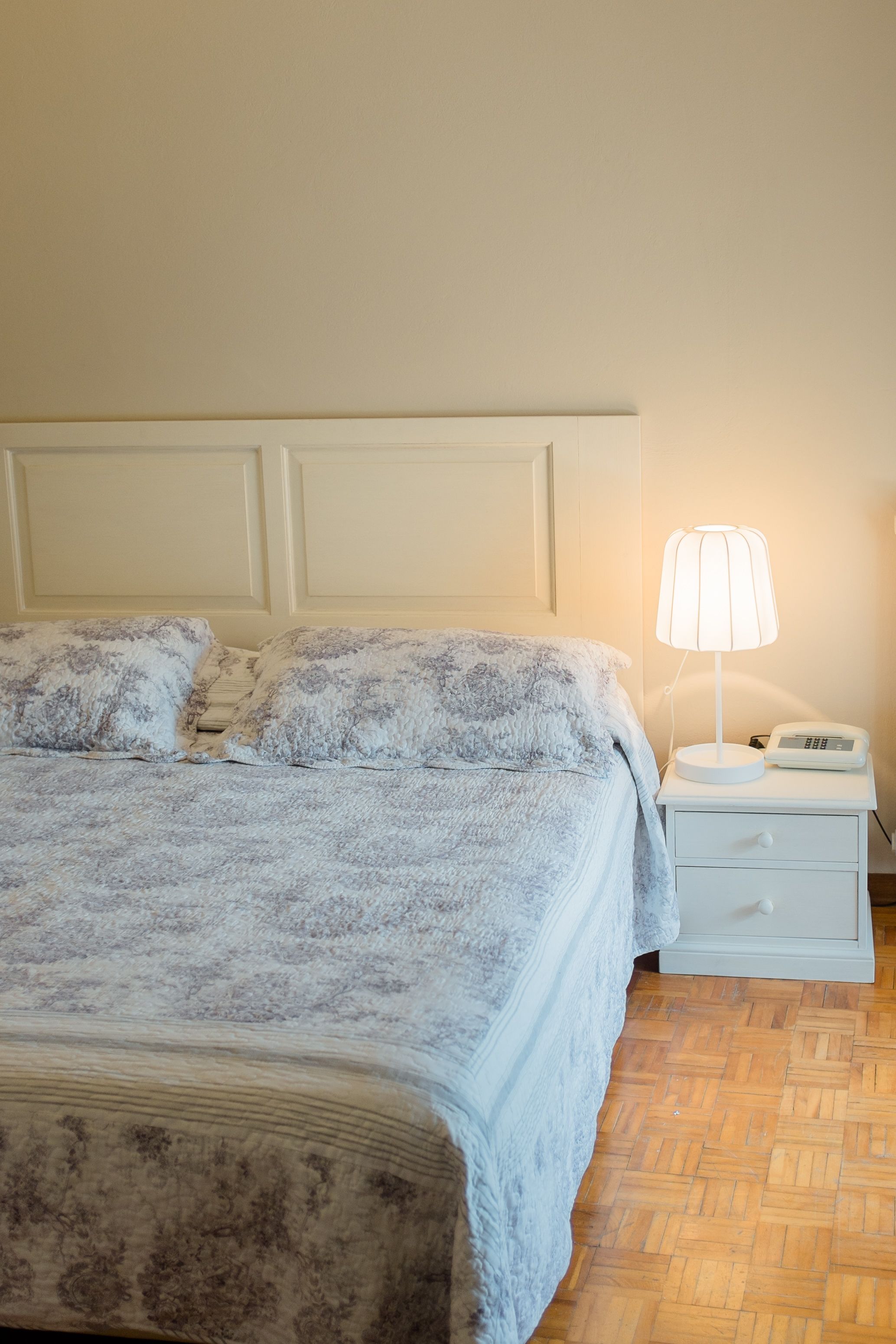 Un letto rifatto con cura, una coperta a motivi floreali, una lampada da comodino e un comodino bianco con un telefono.