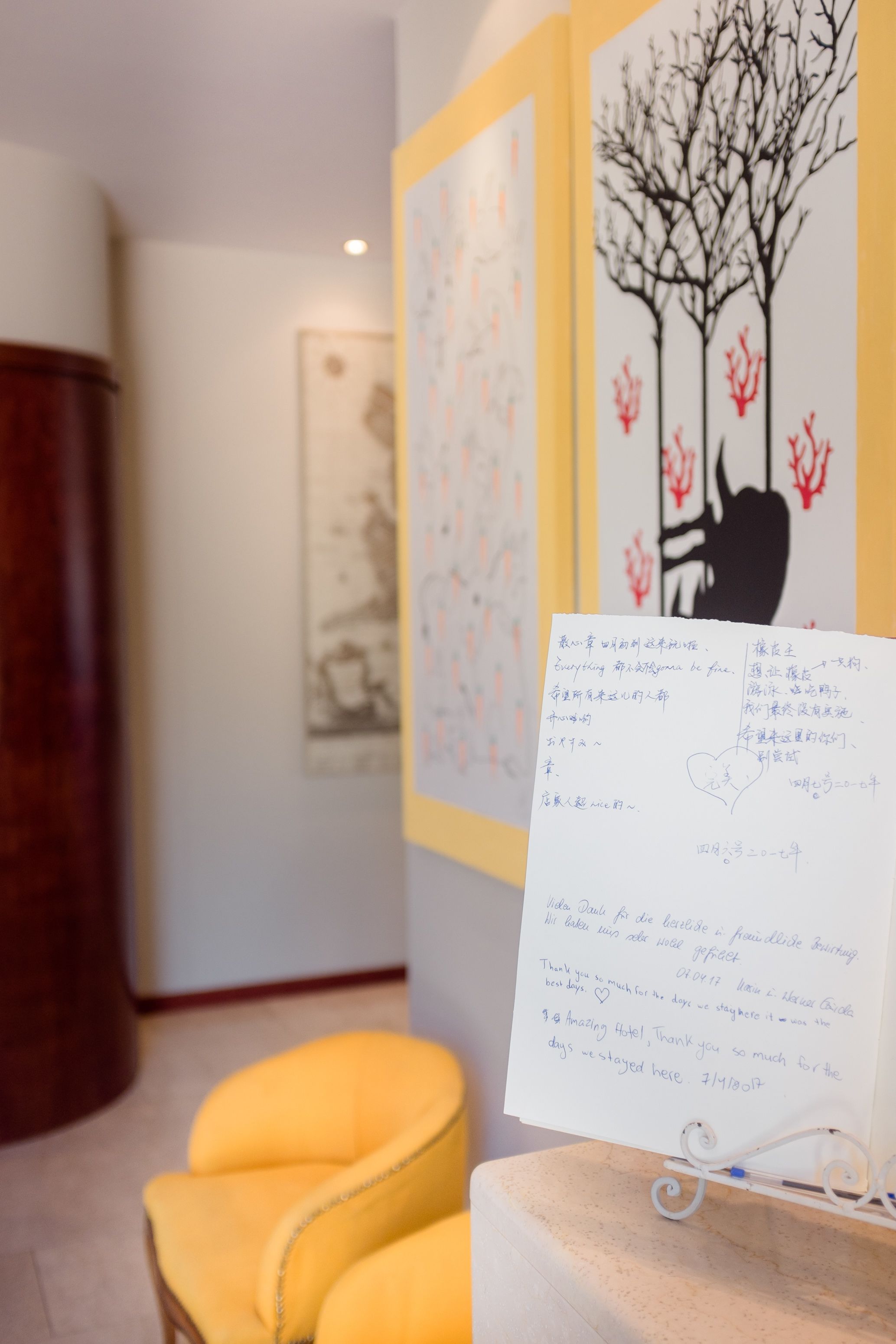 Une pièce avec une chaise jaune, une œuvre d’art encadrée accrochée au mur et un mot écrit à la main posé sur un présentoir.
