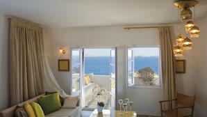A room with a sofa, a coffee table, and a chair. There are curtains and a view of the sea through the windows.