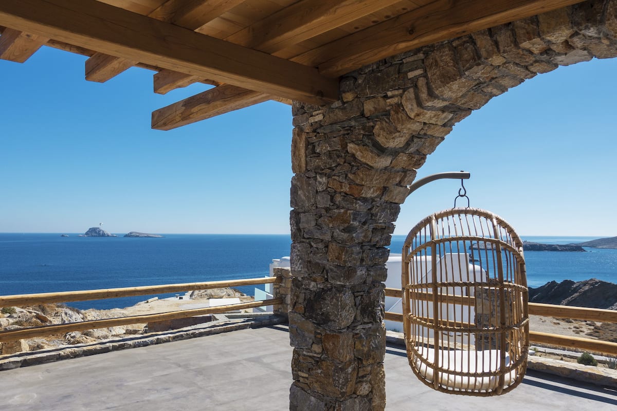 A stone pillar supporting a wooden ceiling, a wicker hanging chair, and a view of the sea with distant islands.