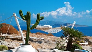 A desert landscape with a white potted cactus, a hammock, and a view of the sea and mountains.