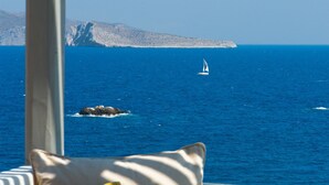 A view from a balcony overlooking a blue sea with a sailboat, rocky islands, and a mountainous landscape.