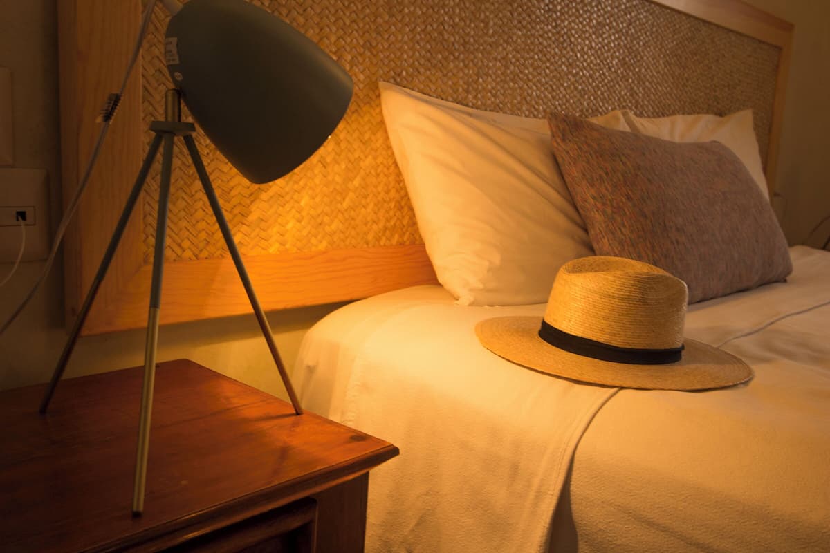 A neatly made bed with a straw hat on the edge, a lamp on a wooden nightstand, and a textured wall behind the bed.