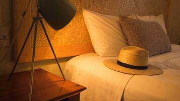 Una cama bien hecha con un sombrero de paja en el borde, una lámpara sobre una mesita de noche de madera y un pared texturizado detrás de la cama.