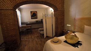 Une chambre d’hôtel avec une arche en briques, un lit agrémenté d’un chapeau de paille et d’un panier, un bureau avec une lampe et un tableau accroché au mur.
