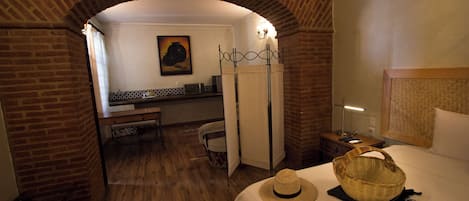 Habitación de hotel con un arco de ladrillo, una cama con sombrero de paja y cesta, un escritorio con lámpara y un cuadro en la pared.
