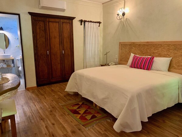 A bedroom with a wooden headboard, a bed with white bedding, a striped pillow, a wooden floor, a small round table, a mirror, a wardrobe, and a wall-mounted light fixture. - Hotel Trebol (Oaxaca)