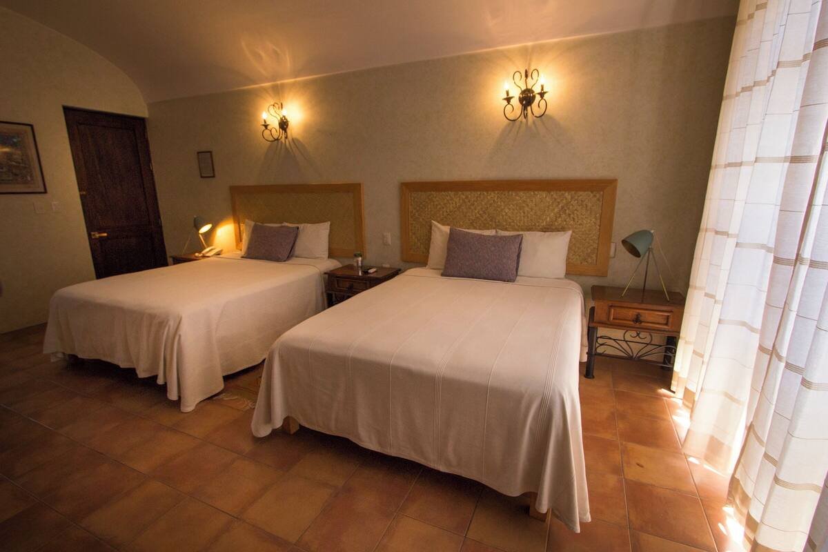 A hotel room with two beds, a wooden headboard, a nightstand, a lamp, and a curtain.