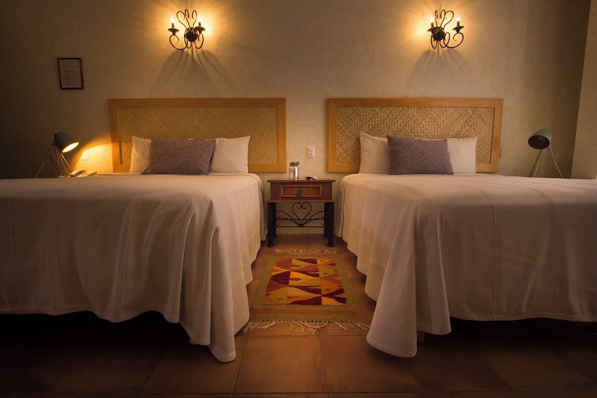 A hotel room with two beds, a small bedside table, a nightstand, a decorative rug, and wall sconces.