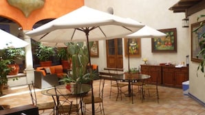 Un espace extérieur aménagé en terrasse, avec une table ronde, des chaises et des parasols. On y trouve également des plantes en pot et un canapé avec des coussins orange.