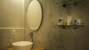 Un baño con pared de mármol, espejo redondo, tocador de madera, lavamanos blanco y un inodoro.