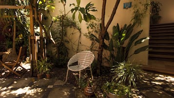 Uma cadeira de madeira em área externa sombreada, com plantas em vasos e piso de azulejos.