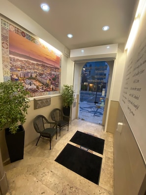 A hotel lobby with a large cityscape mural, two potted plants, a reception desk, and a black area rug.