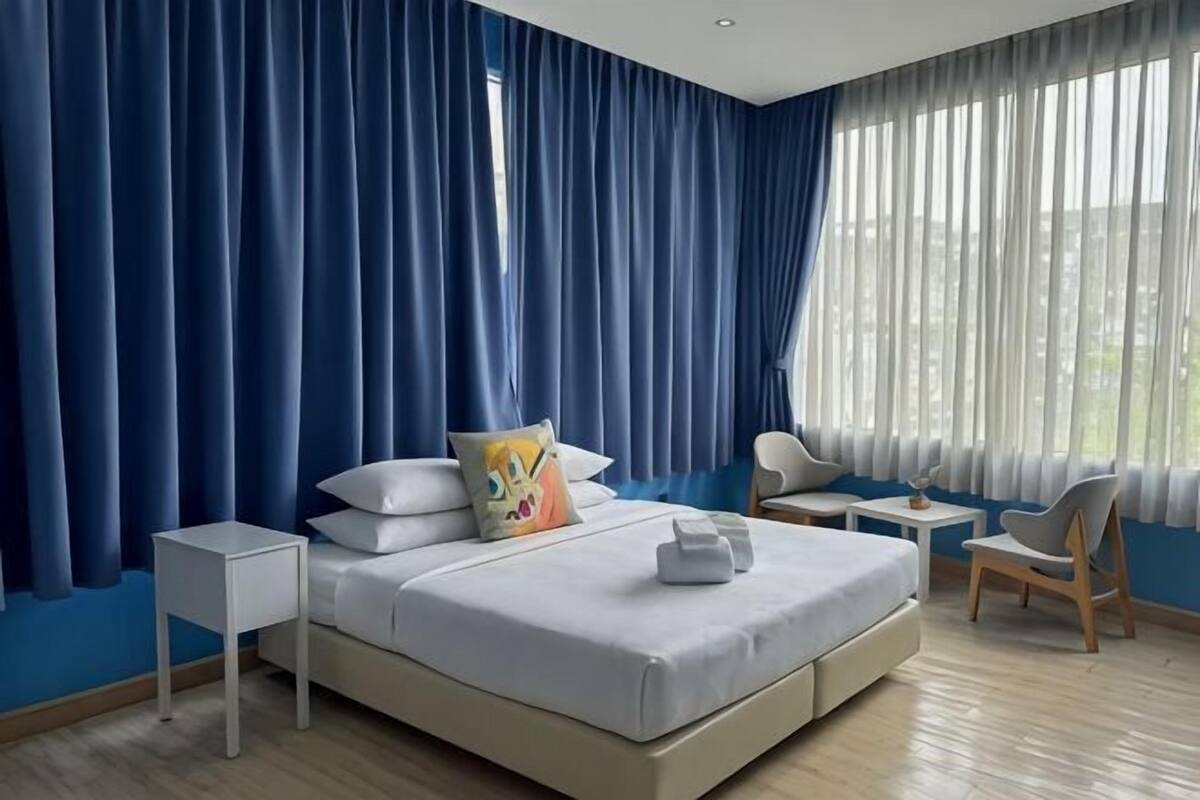 Une chambre d’hôtel moderne, dotée d’un grand lit, d’une table de chevet, d’une chaise et offrant une vue sur la ville grâce à de grandes fenêtres.