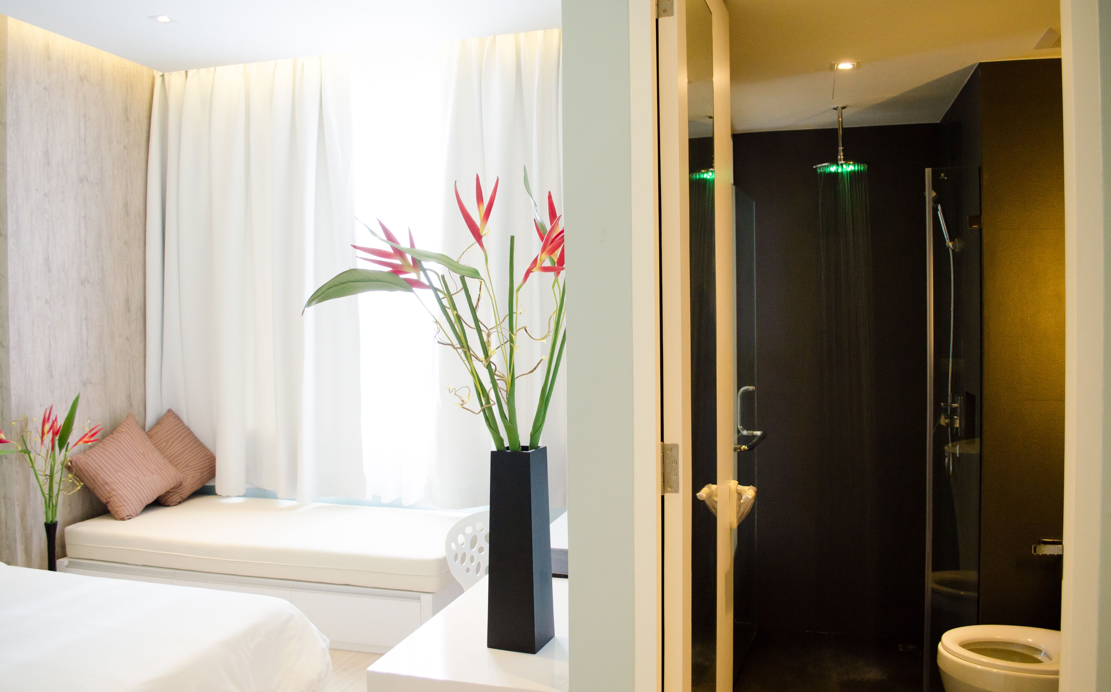A modern hotel room with a bed, a bedside table with a vase of flowers, a window with curtains, and a view of a shower area.