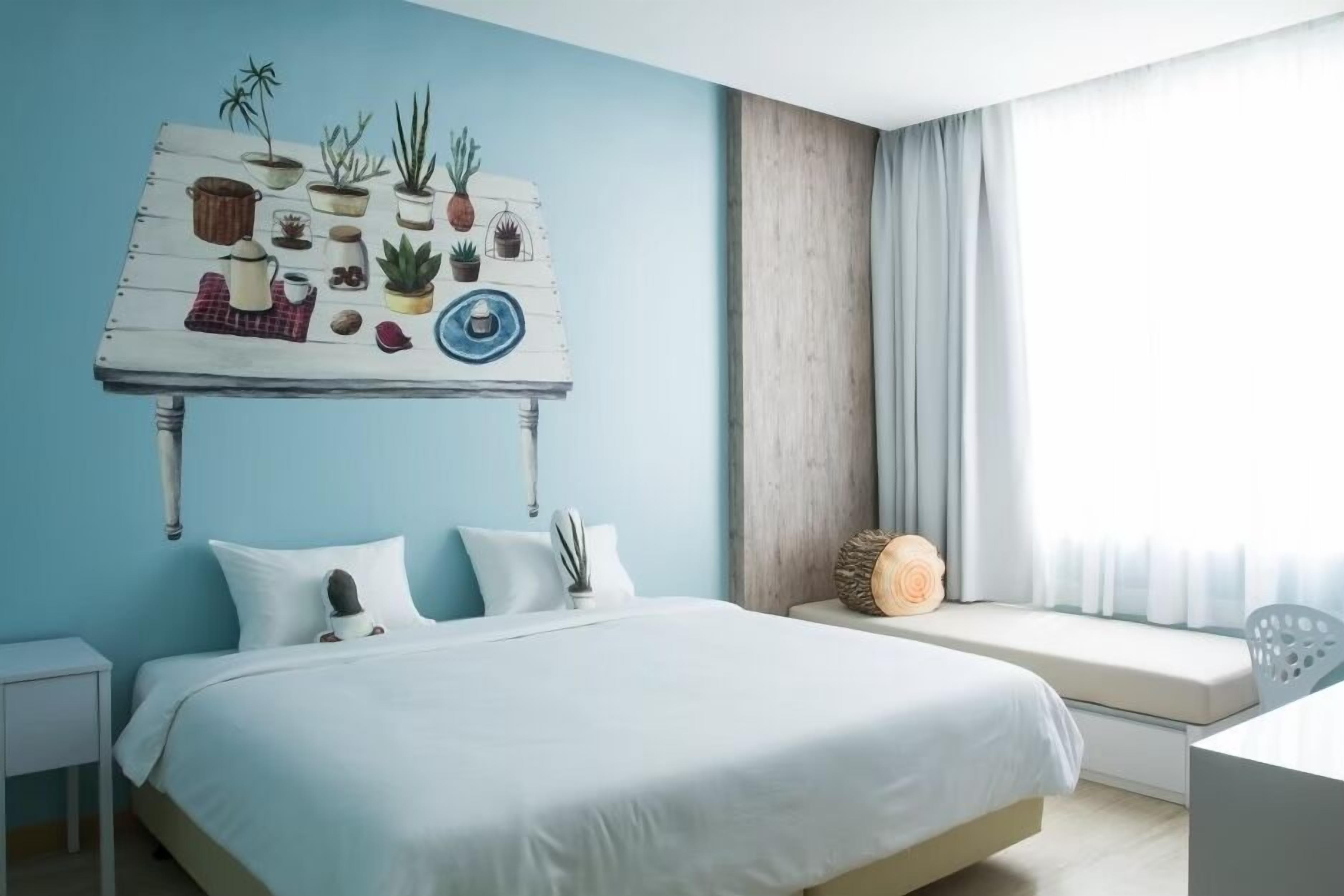 Une chambre avec un grand lit, une table de chevet, un coin fenêtre aménagé et une œuvre d’art murale représentant des plantes en pot et un œil bleu.