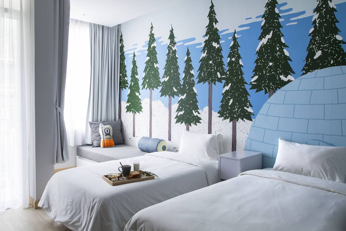 Une chambre d’hôtel avec deux lits, une fresque représentant des arbres, une fenêtre avec des rideaux et une table de chevet sur laquelle se trouve un plateau avec des articles pour le petit-déjeuner.