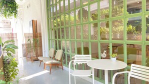 A sunlit patio with a white table and chairs, large green-framed windows, and hanging plants. - Beat Hotel Bangkok (Bangkok)